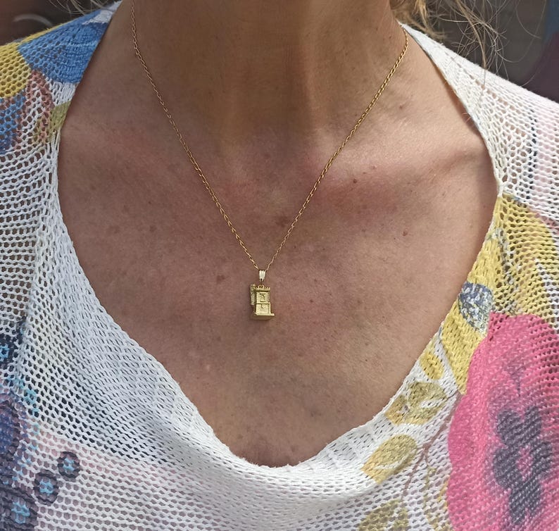 Op de afbeelding: Een gouden ketting met een klein gouden hanger in de vorm van een gebouw. De hanger heeft een gedetailleerd ontwerp en hangt aan de borst van een vrouw.
