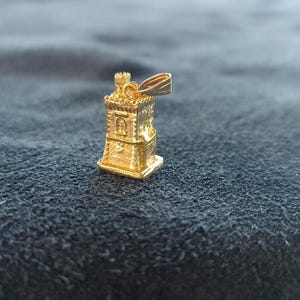 Op de afbeelding: Een gouden hanger in de vorm van een toren met een vlag erop. De hanger is gedetailleerd met ramen en een dak.