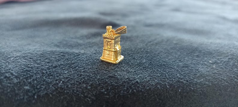 Op de afbeelding: Een gouden hanger in de vorm van een toren met een vlag erop. De hanger is gedetailleerd met ramen en een dak.