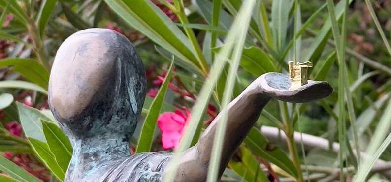 Op de afbeelding: Een bronzen beeld van een persoon met een uitgestrekte arm die een klein gouden model van een gebouw in zijn handpalm houdt.