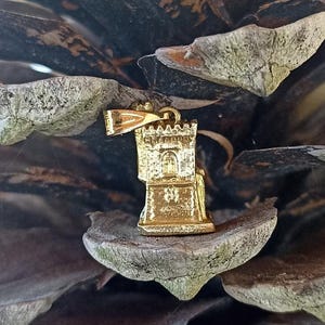 Op de afbeelding: Gouden metalen hanger in de vorm van een toren met een klein raam en een decoratieve bovenkant. De hanger hangt aan een dennenappel.