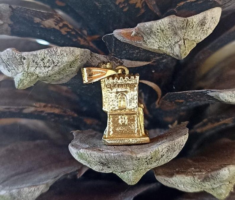 Op de afbeelding: Gouden metalen hanger in de vorm van een toren met een klein raam en een decoratieve bovenkant. De hanger hangt aan een dennenappel.