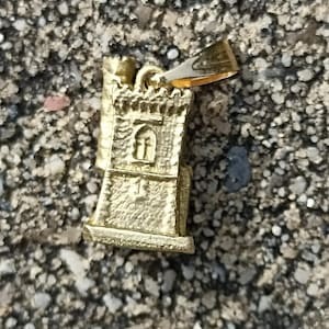 Op de afbeelding: Gouden metalen hanger in de vorm van een kleine toren met een raam en een decoratieve bovenkant. De hanger heeft een lus aan de bovenkant om aan een ketting of armband te bevestigen.