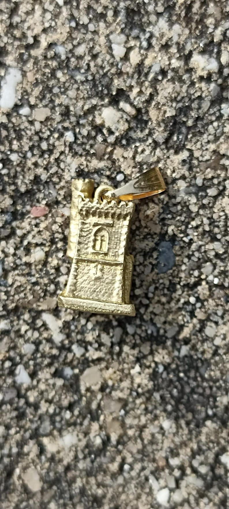 Op de afbeelding: Gouden metalen hanger in de vorm van een kleine toren met een raam en een decoratieve bovenkant. De hanger heeft een lus aan de bovenkant om aan een ketting of armband te bevestigen.