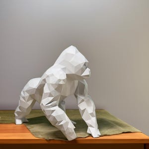 Puede incluir: Escultura de gorila geométrico blanco con un diseño de bajo polígono. La escultura está de pie sobre una superficie de madera con una tela verde debajo.