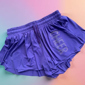 Puede incluir: Pantalones cortos de animadora morados con un diseño en capas. Los pantalones cortos tienen una cintura elástica con cordón y la palabra "CHEER" con pedrería.