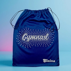 May include: A royal blue drawstring bag with the word "Gymnast" spelled out in silver rhinestones. The name "Elaina" is also spelled out in silver rhinestones below the word "Gymnast". The bag has black drawstrings.