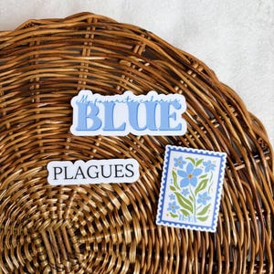 May include: Two stickers on a woven wicker surface. One sticker says "My favorite color is BLUE" in blue text. The other sticker is a blue and green stamp with a floral design and the text "HEYA".