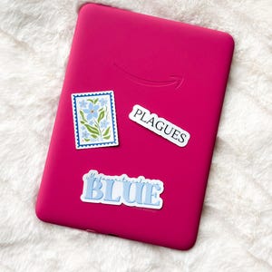 May include: A pink e-reader with three stickers on it. The stickers are a floral design, a sticker that says "My favorite color is BLUE", and a sticker that says "PLAGUES".