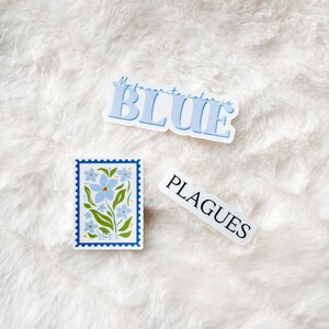 May include: Three stickers on a white fuzzy surface. One sticker says "My favorite color is BLUE" in blue letters. Another sticker has a blue flower design with the word "HEXA" at the bottom. The third sticker says "PLAGUES" in white letters.