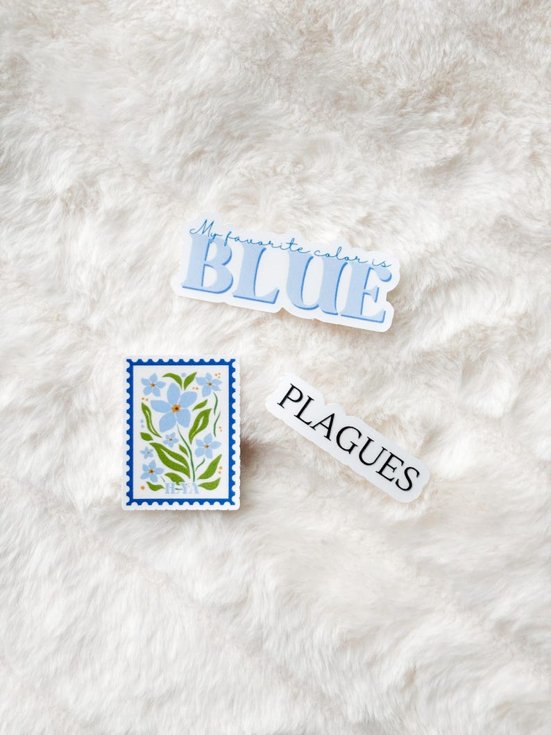 May include: Three stickers on a white fuzzy surface. One sticker says "My favorite color is BLUE" in blue letters. Another sticker has a blue flower design with the word "HEXA" at the bottom. The third sticker says "PLAGUES" in white letters.