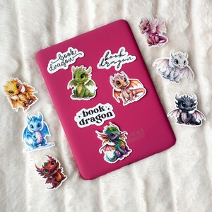 May include: A pink laptop with nine colorful dragon stickers. The stickers feature different colored dragons with the text "book dragon" on three of the stickers.