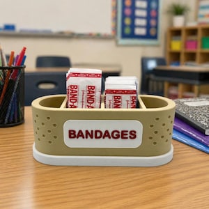 May include: A beige and white bandage holder with the word "BANDAGES" in red lettering. The holder is filled with individually wrapped bandages. The holder is on a wooden desk in a classroom setting.