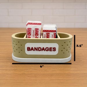 May include: A beige and white rectangular bandage holder with the word "BANDAGES" in red. The holder is 6 inches long and 2.5 inches tall, and contains several individually wrapped bandages.