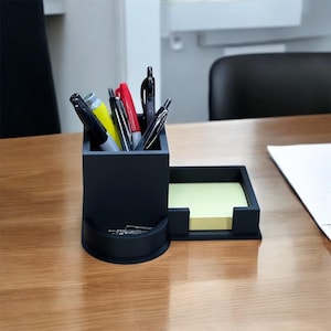 May include: Black desk organizer with a pen holder, a note pad holder, and a small compartment for paper clips. The organizer is on a wooden desk.