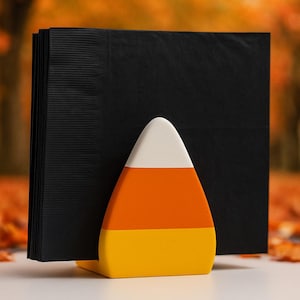 May include: A Halloween-themed napkin holder with a candy corn design. The holder is shaped like a candy corn with white, orange, and yellow sections. Black napkins are held in place by the candy corn-shaped holder.