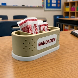 May include: A beige and white rectangular container with the word "BANDAGES" in red lettering. The container is filled with individually wrapped adhesive bandages. The container is on a wooden desk in a classroom setting.