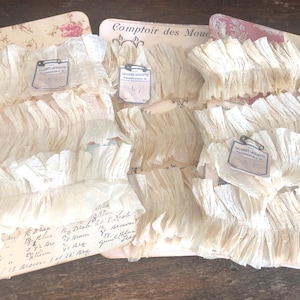 May include: Three packages of white ruffled trim, each with a small tag attached. The tags are labeled "Comtoir des Mouchoirs" and have a handwritten number on them. The trim is made of a delicate, crinkled fabric and is perfect for adding a touch of vintage charm to any project.