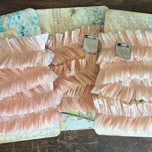 May include: Several stacks of pale pink ruffled trim are displayed on decorative cards with vintage-style designs. Each trim stack has a small tag attached. The cards feature floral and script patterns, with a butterfly and bamboo illustration.