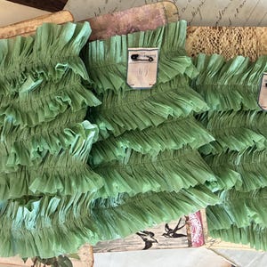 May include: Three sage green ruffled fabric strips arranged side by side. Each strip features a small white rectangular tag attached with a black safety pin. The tags display the text Galeries Lafayette in black ink. The fabric has a pleated texture.