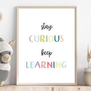 May include: A framed print with the text "stay curious keep learning" in colorful letters on a white background. The print is hanging on a white wall with a wooden shelf below it.