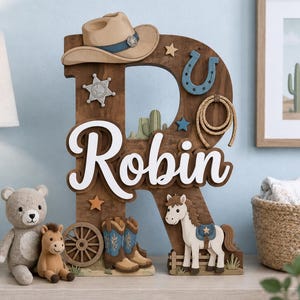Può includere: Una grande lettera R in legno a tema western con il nome Robin in corsivo bianco. Le decorazioni includono un cappello da cowboy beige, una stella da sceriffo argentata, un ferro di cavallo blu, una corda, una ruota e un cavallo.