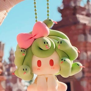 May include: A quirky keychain depicting a cartoon character with a light green snake-like hairstyle and a pink bow. The character has a light skin tone, red eyes, and a cute expression. The snakes have red tongues.