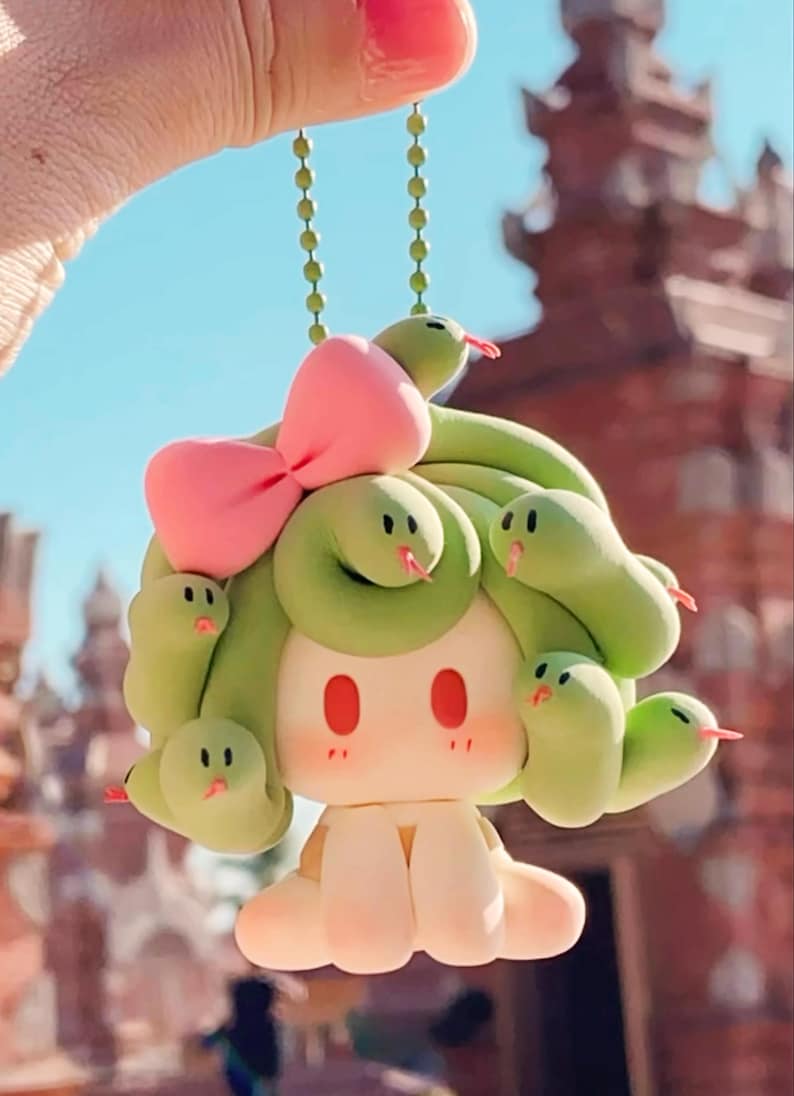 May include: A quirky keychain depicting a cartoon character with a light green snake-like hairstyle and a pink bow. The character has a light skin tone, red eyes, and a cute expression. The snakes have red tongues.