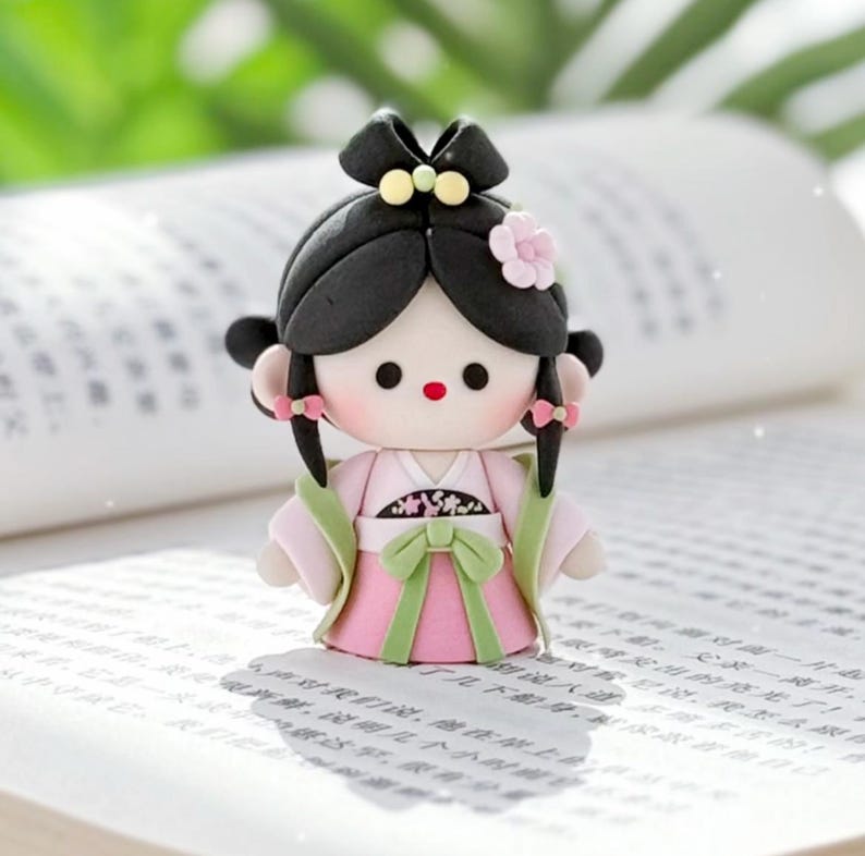 May include: A handmade doll figurine of a woman in traditional Chinese dress. The doll has a pink dress with green details, black hair, and a flower hair accessory. The doll is placed on an open book.
