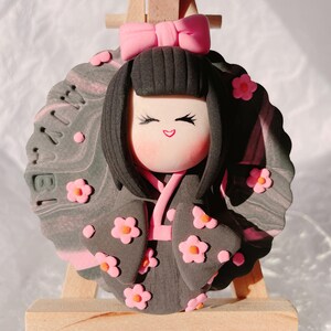 May include: A handcrafted doll with a smiling face, black hair, and a pink bow. The doll wears a dark grey kimono adorned with pink flower accents. The background is a grey and pink marbled design with the number "18" visible.