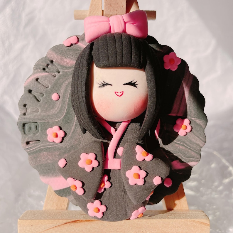 May include: A handcrafted doll with a smiling face, black hair, and a pink bow. The doll wears a dark grey kimono adorned with pink flower accents. The background is a grey and pink marbled design with the number "18" visible.