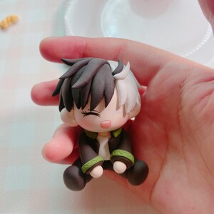 May include: A handmade figurine of a seated character with black and white hair, wearing a dark jacket with green trim. The figurine has closed eyes and a gentle expression, with a light skin tone and rosy cheeks. The figure is held in a hand.
