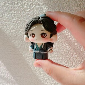 May include: A miniature figurine of a character with dark hair and red eyes, dressed in a dark blue outfit with teal details. The figure has a cartoonish style and is held in a person's hand.