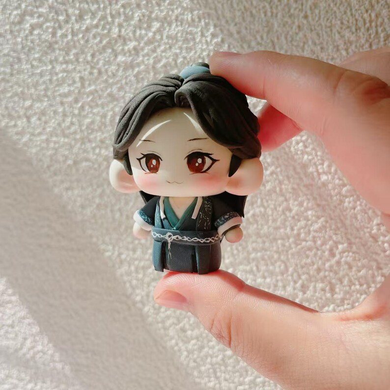 May include: A miniature figurine of a character with dark hair and red eyes, dressed in a dark blue outfit with teal details. The figure has a cartoonish style and is held in a person's hand.