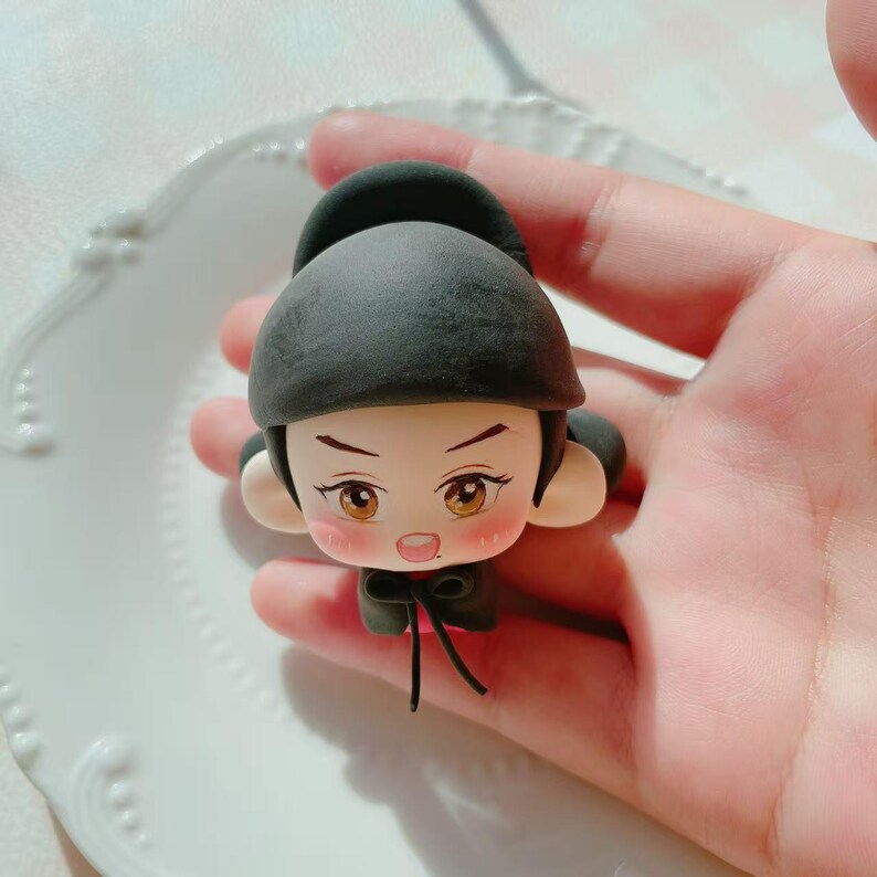 May include: A miniature figurine of a person wearing a black hat and outfit. The figure has a detailed face with brown eyes and rosy cheeks. The figurine is held in a hand, highlighting the craftsmanship of the small sculpture. The figurine is likely made of clay.