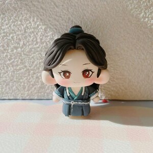 May include: A handmade figurine of a character with dark hair, red eyes, and rosy cheeks. The figure is dressed in a blue and teal outfit with detailed features. The doll has a calm expression and is standing upright.