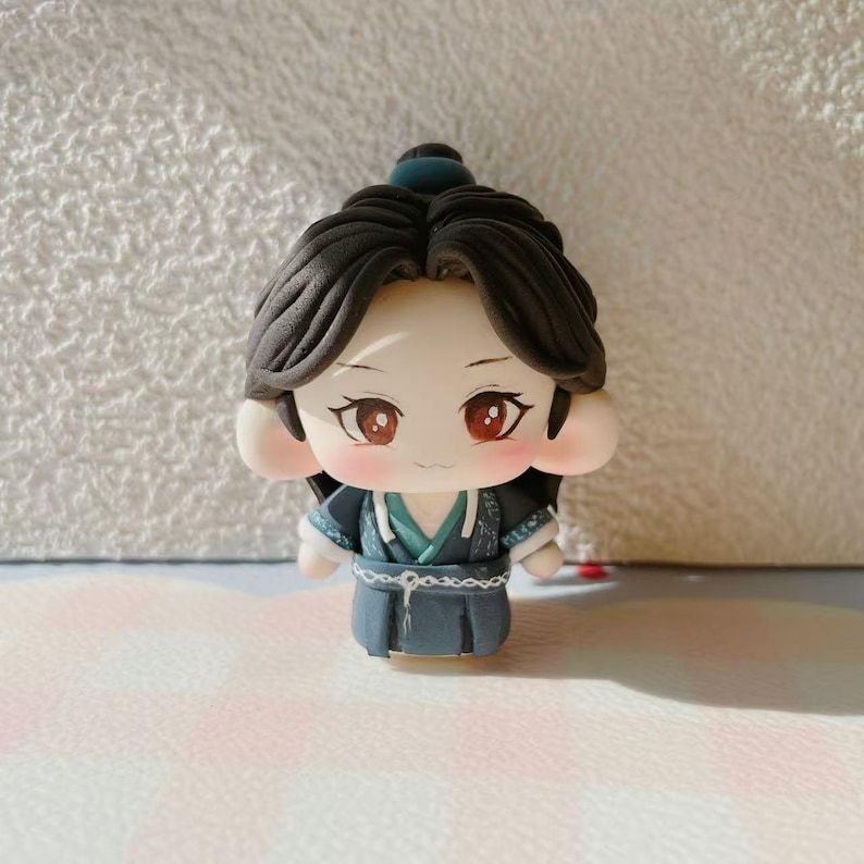May include: A handmade figurine of a character with dark hair, red eyes, and rosy cheeks. The figure is dressed in a blue and teal outfit with detailed features. The doll has a calm expression and is standing upright.