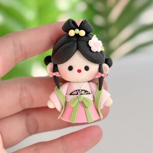 May include: A handmade doll figurine in traditional Asian dress. The doll has black hair styled in a bun with a flower and yellow details. The doll wears a pink dress with green accents and a green bow.