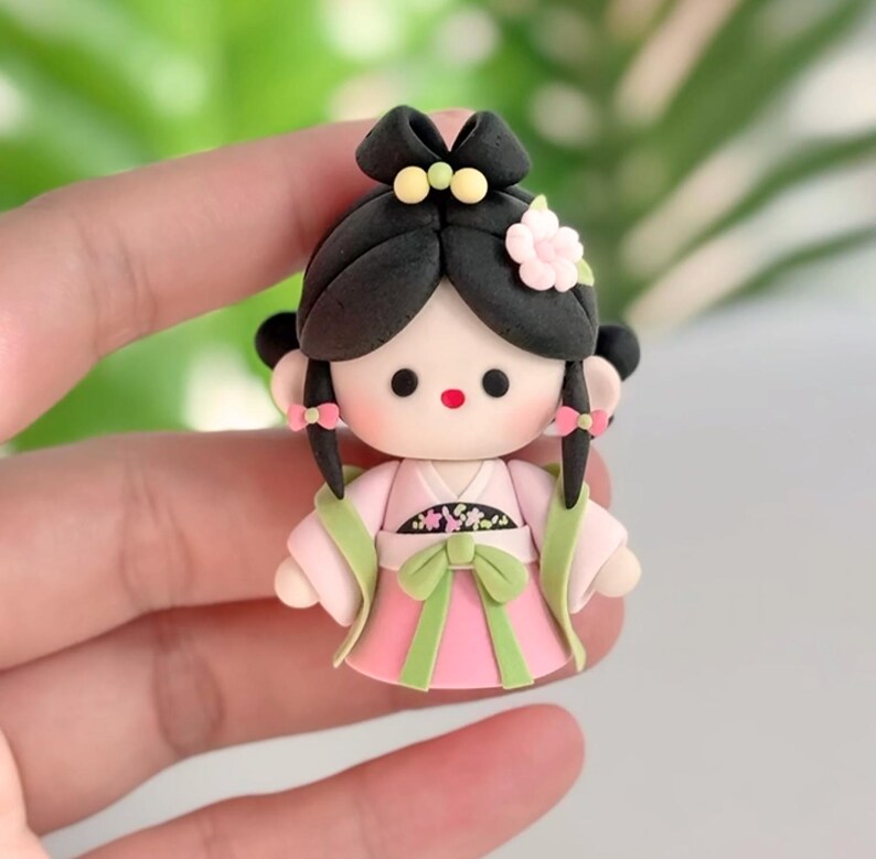 May include: A handmade doll figurine in traditional Asian dress. The doll has black hair styled in a bun with a flower and yellow details. The doll wears a pink dress with green accents and a green bow.