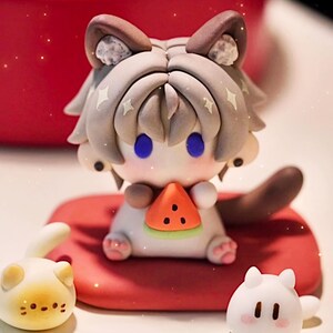 May include: A charming cat-like figurine with a watermelon slice, seated on a red cushion. The figure has grey and brown details, blue eyes, and star accents. Two smaller cat figurines are also present, one yellow and one white.