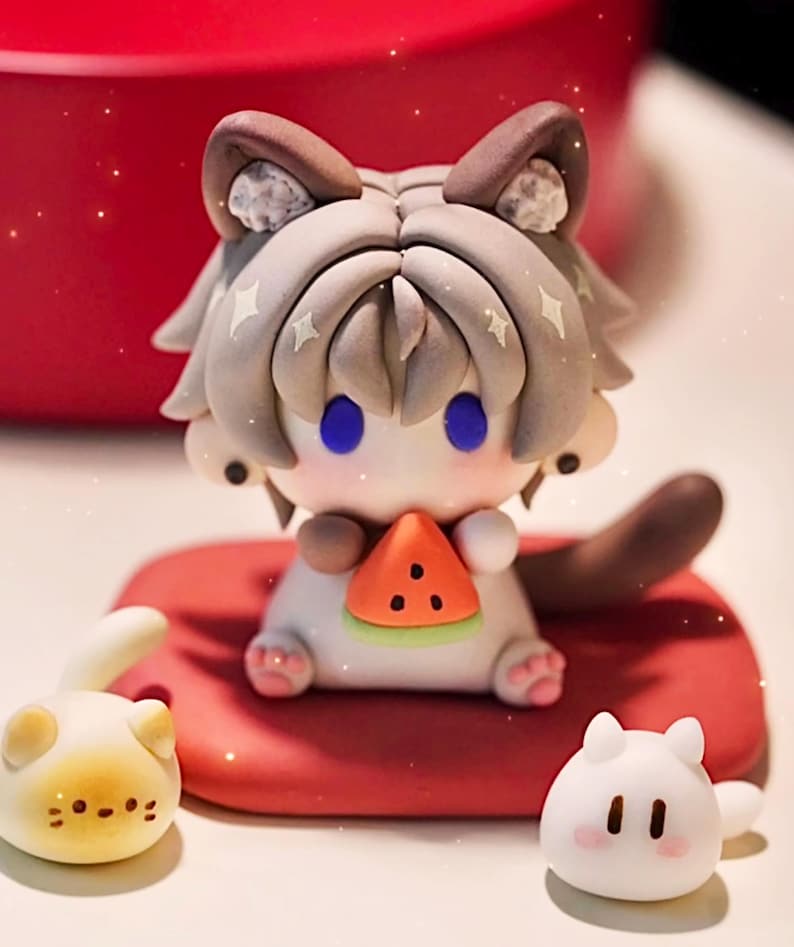 May include: A charming cat-like figurine with a watermelon slice, seated on a red cushion. The figure has grey and brown details, blue eyes, and star accents. Two smaller cat figurines are also present, one yellow and one white.