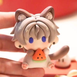 May include: A small, handcrafted figurine of a cat-like character holding a watermelon slice. The figure has grey hair, blue eyes, and brown ears. The watermelon is orange with black seeds. The figurine is white and tan with pink paws.