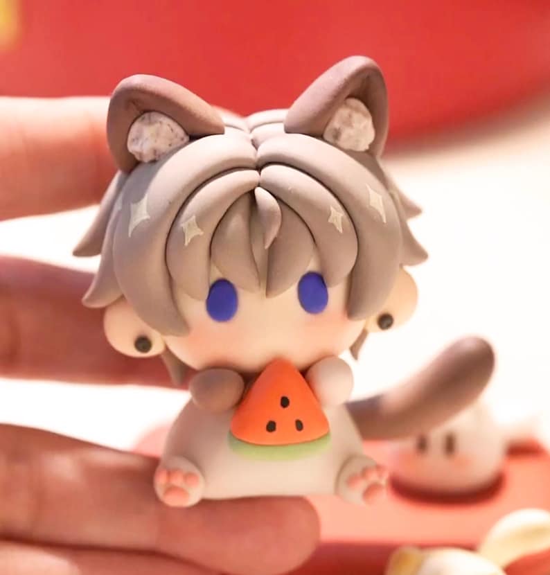 May include: A small, handcrafted figurine of a cat-like character holding a watermelon slice. The figure has grey hair, blue eyes, and brown ears. The watermelon is orange with black seeds. The figurine is white and tan with pink paws.