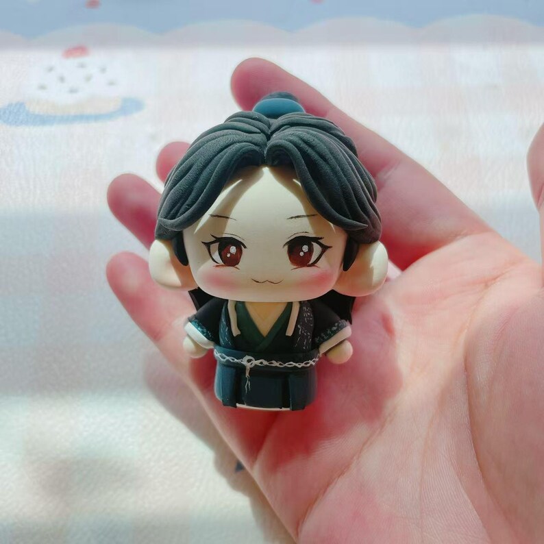 May include: A miniature figurine of a character with dark hair and a traditional outfit. The figure has large, expressive eyes and a gentle expression. The outfit is primarily dark with green and white accents. The figurine is held in a hand.