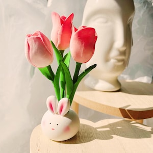 Pode incluir: Um vaso de cerâmica caprichoso em forma de coelho, segurando três flores de tulipa rosa com caules e folhas verdes. O vaso de coelho tem bochechas rosadas e orelhas forradas de rosa. Um busto branco está no fundo.