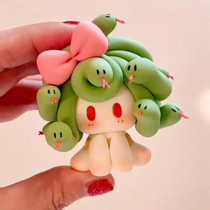May include: A charming clay figurine featuring a character with green snake-like hair, a pink bow, and red eyes. The figure has a pale complexion and a small, smiling face. The snakes have pink tongues.