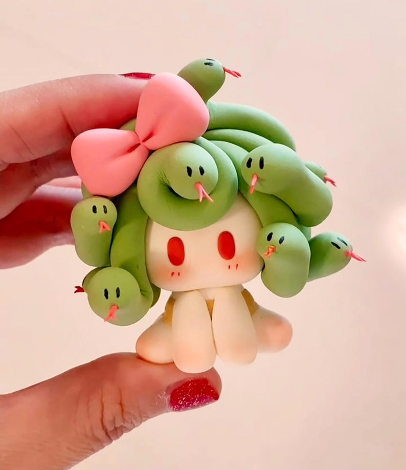 May include: A charming clay figurine featuring a character with green snake-like hair, a pink bow, and red eyes. The figure has a pale complexion and a small, smiling face. The snakes have pink tongues.