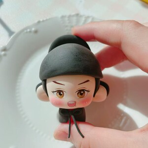 May include: A handmade figurine of a cartoon character wearing a black hat and outfit. The character has a light skin tone, rosy cheeks, and large brown eyes. The figurine is held by a person's hand.