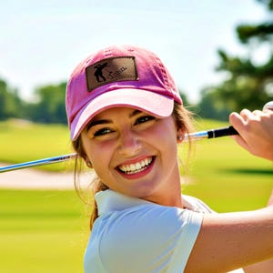 Pode incluir: Uma mulher com boné de golfe rosa com logotipo, camisa branca e luva de golfe, balança um taco de golfe em um campo ensolarado. O fundo apresenta grama verde e árvores.