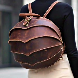 Könnte beinhalten: Ein brauner Lederrucksack mit einem einzigartigen, geschichteten Design. Der Rucksack hat eine abgerundete Form mit mehreren geschwungenen Abschnitten und weißen Nähten. Er verfügt über verstellbare Träger und einen oberen Griff, der mit einem goldfarbenen Verschluss befestigt ist.